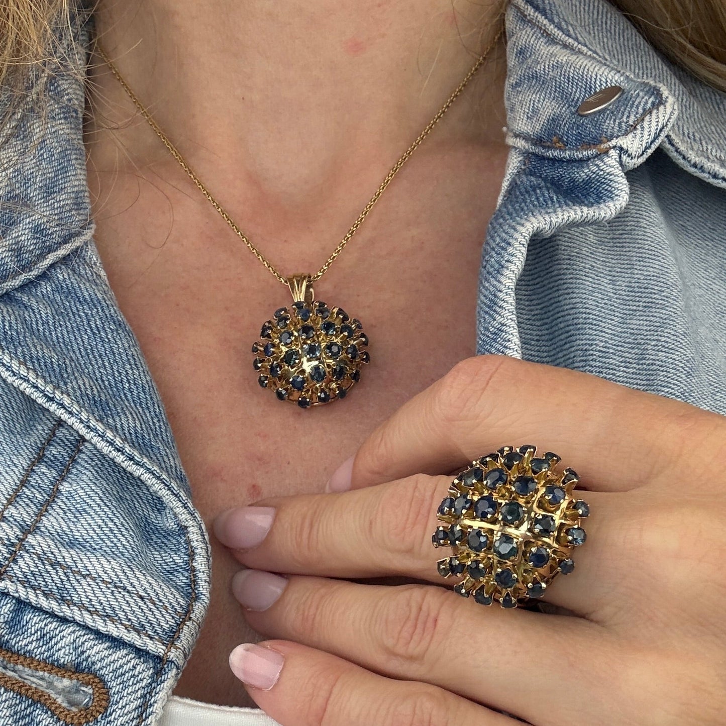 Close-up of hands wearing a gold sapphire pendant necklace and gold sapphire ring with a denim jacket background