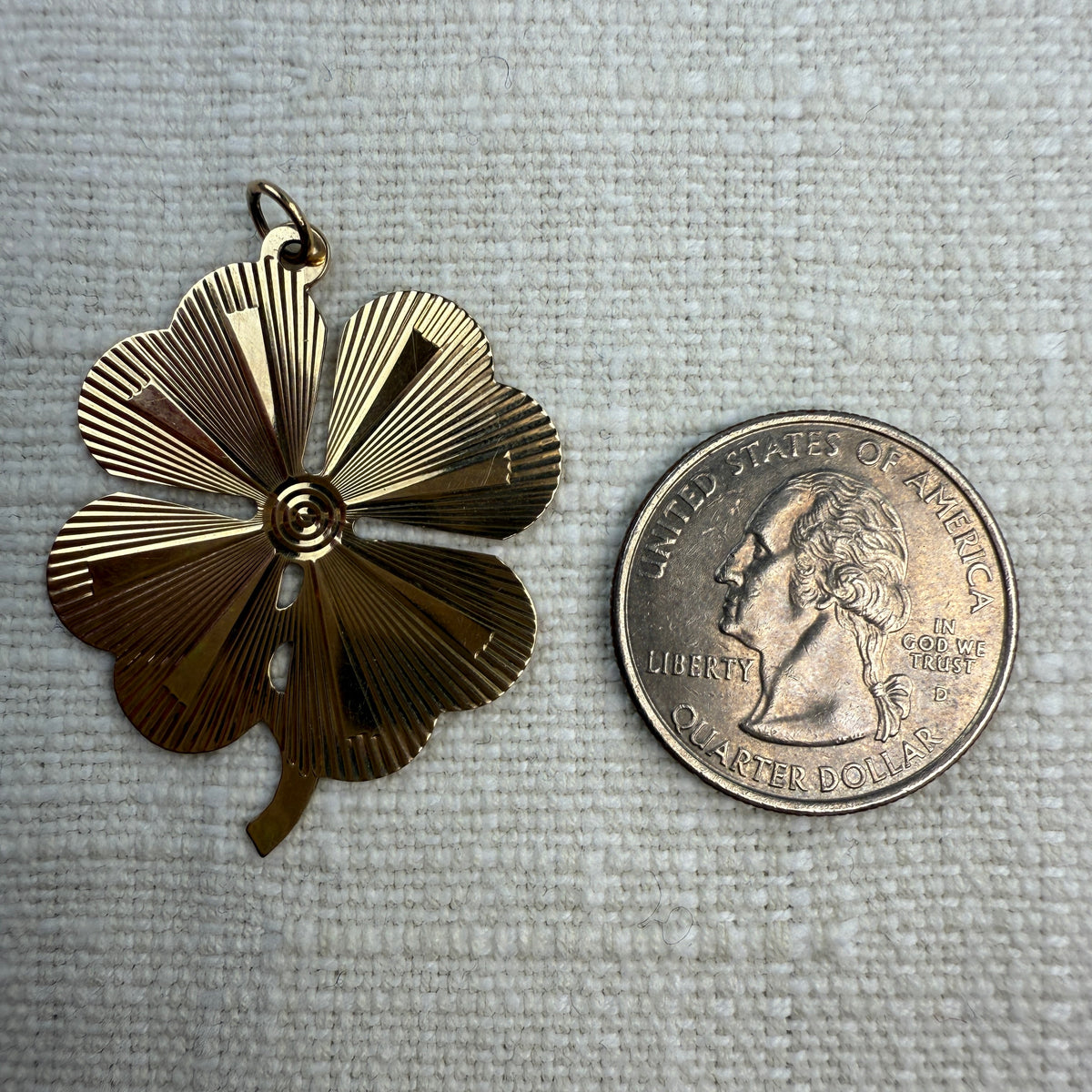 Engine turned vintage four-leaf clover pendant on an off white linen background next to a quarter. Showing that it is larger than a quarter.