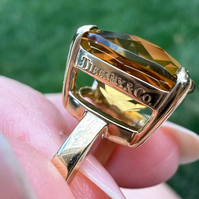 A gold ring with a cushion-cut yellow gemstone held by fingers. The ring is engraved with 'TIFFANY & CO.