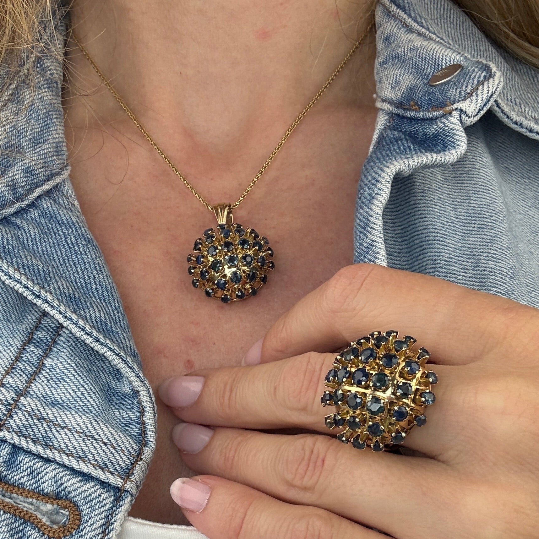 Close-up of hands wearing a gold sapphire pendant necklace and gold sapphire ring with a denim jacket background