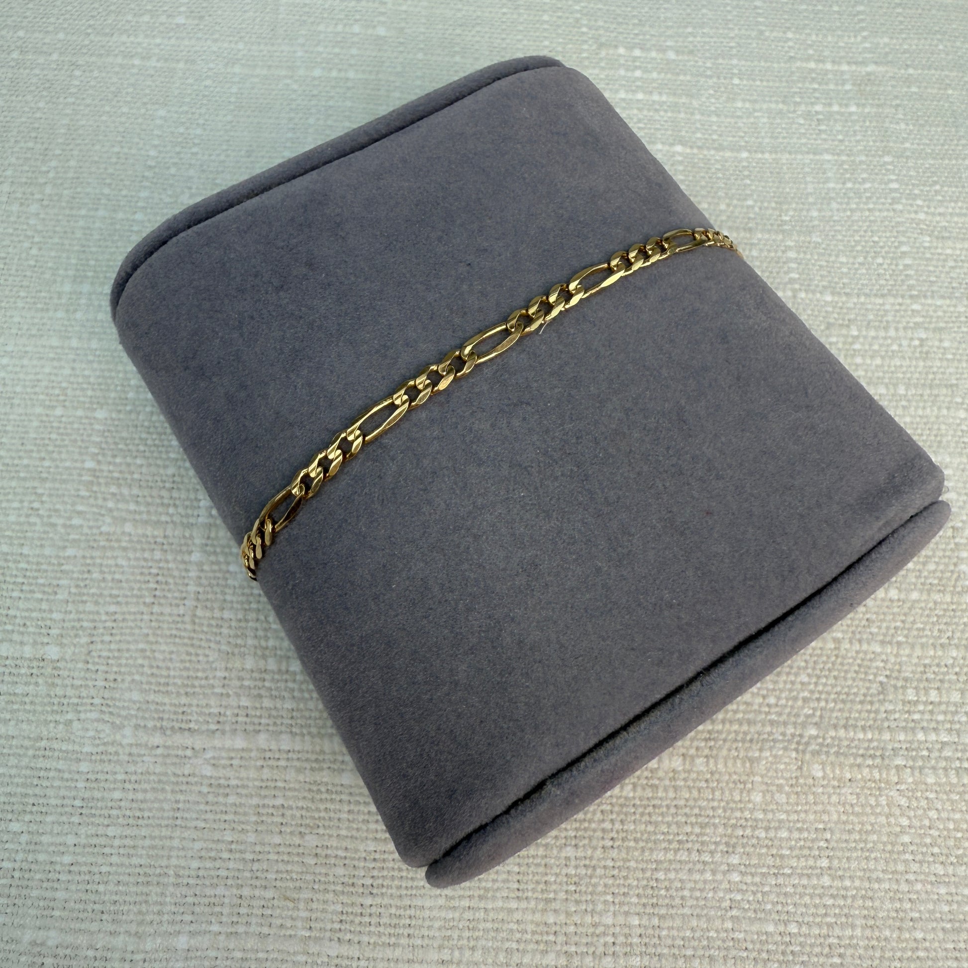 Gold figaro link bracelet displayed on a gray fabric pillow.