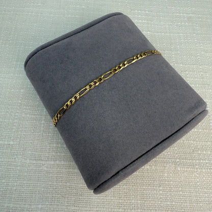 Gold figaro link bracelet displayed on a gray fabric pillow.