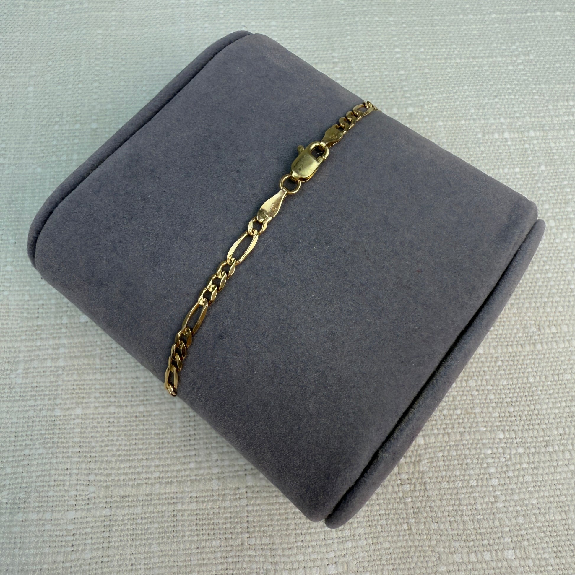Gold figaro link bracelet displayed on a gray fabric pillow, highlighting the lobster clasp.