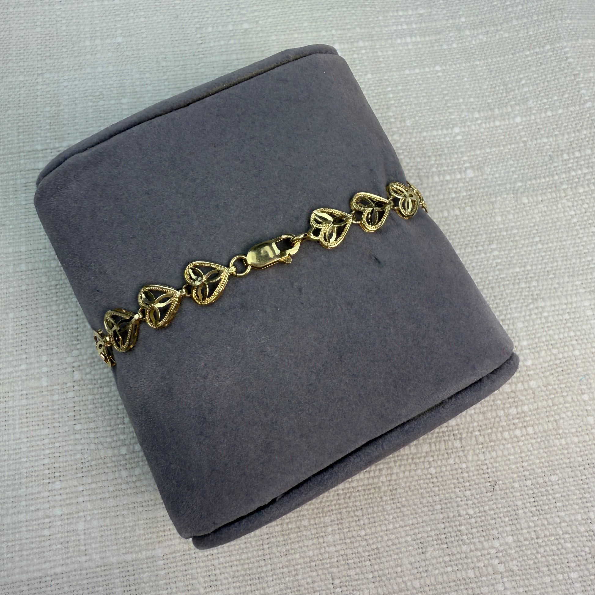 Gold filigree heart bracelet displayed on a gray fabric pillow, highlighting a lobster clasp.