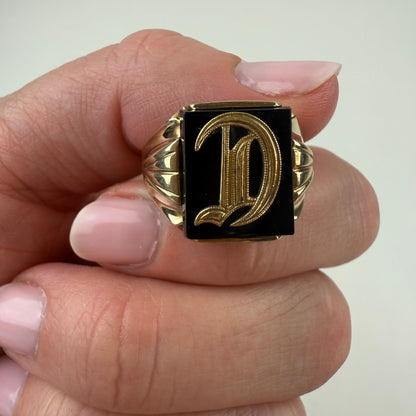 Gold ring with a black square and letter 'D' held by a hand against a white background