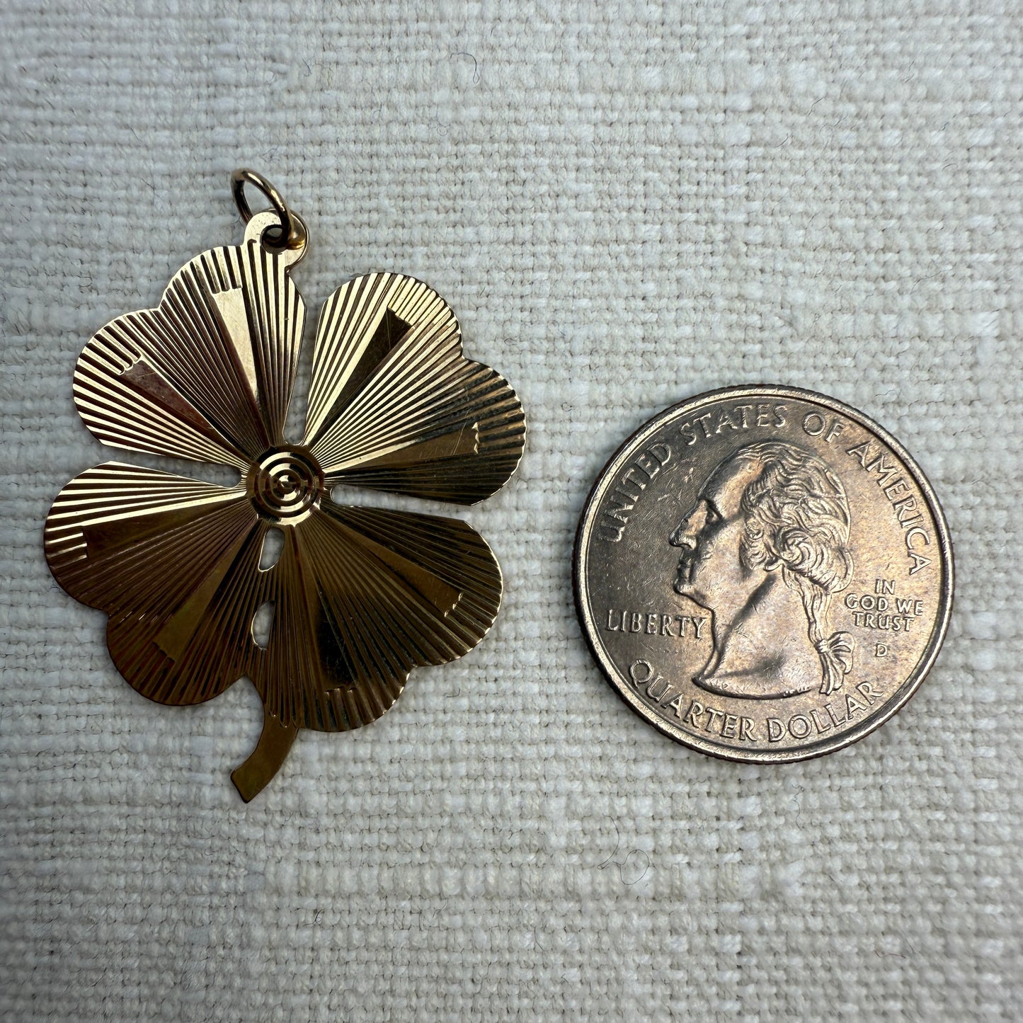 Engine turned vintage four-leaf clover pendant on an off white linen background next to a quarter. Showing that it is larger than a quarter. 