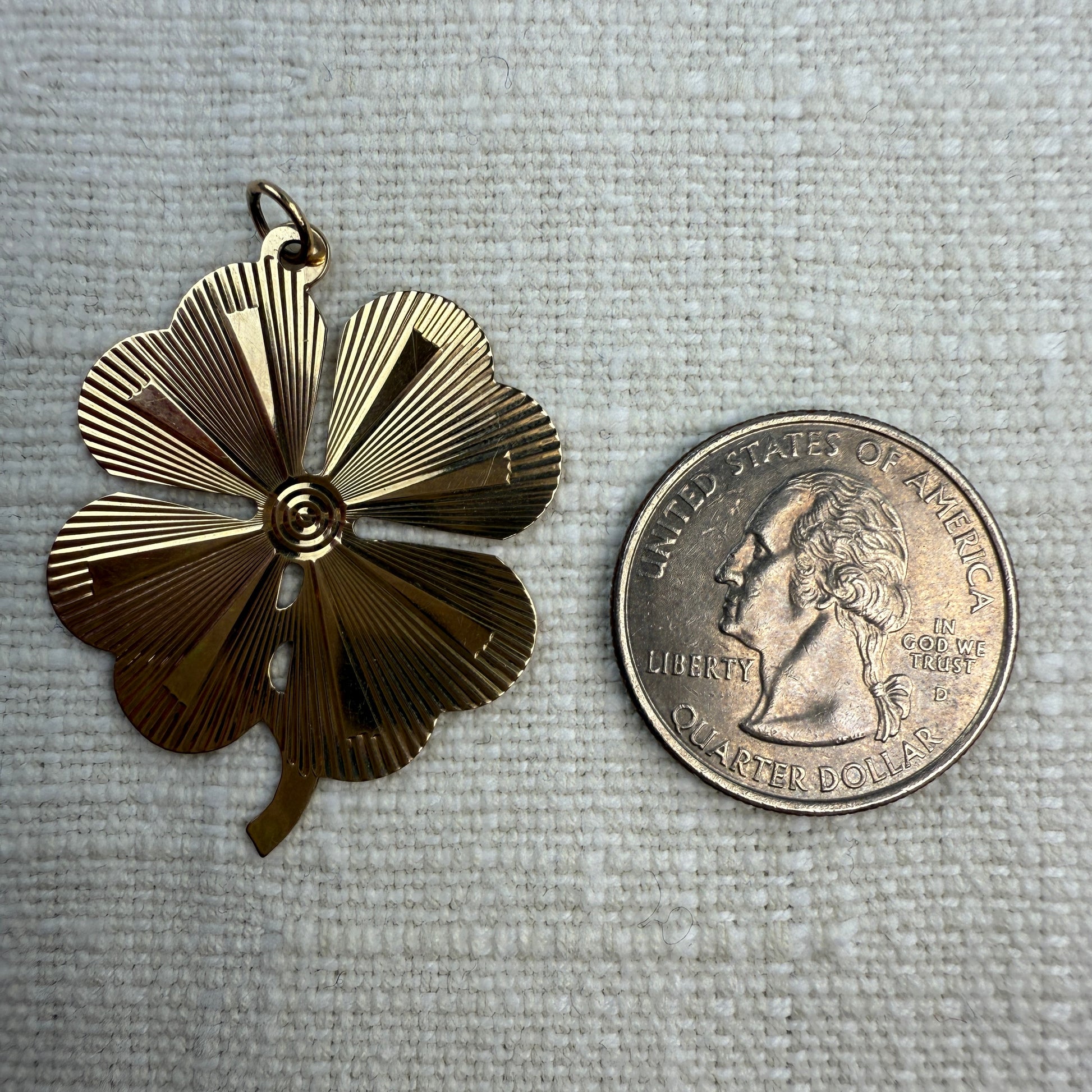 Engine turned vintage four-leaf clover pendant on an off white linen background next to a quarter. Showing that it is larger than a quarter. 