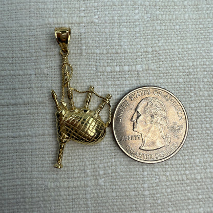 Gold bagpipe pendant next to a U.S. quarter dollar on a gray fabric background
