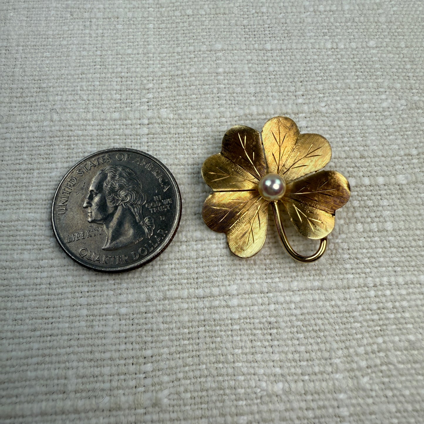 Gold four-leaf clover brooch  and pendant with a pearl next to a U.S. quarter on a textured gray background