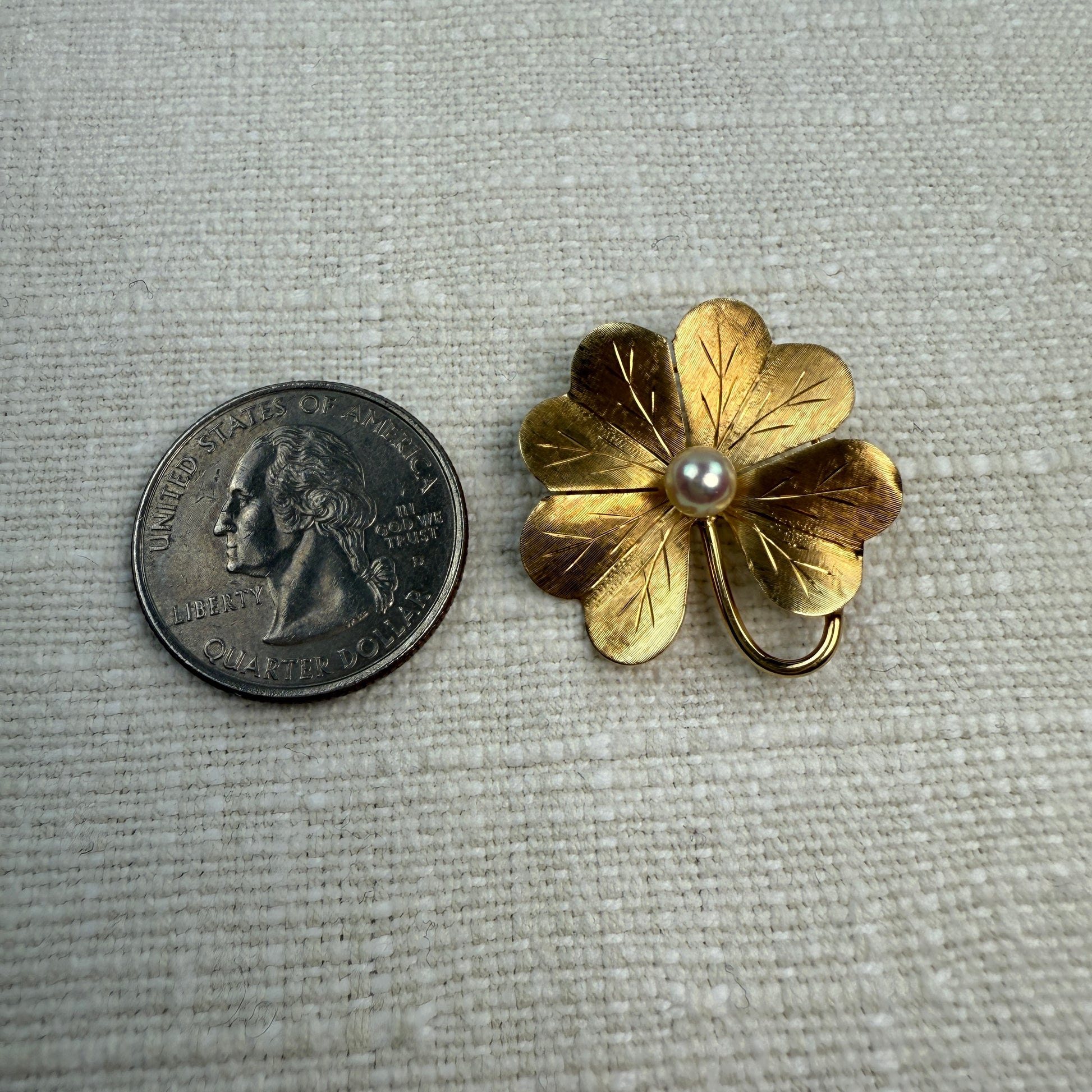 Gold four-leaf clover brooch  and pendant with a pearl next to a U.S. quarter on a textured gray background