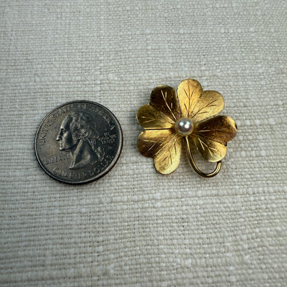Gold four-leaf clover brooch  and pendant with a pearl next to a U.S. quarter on a textured gray background
