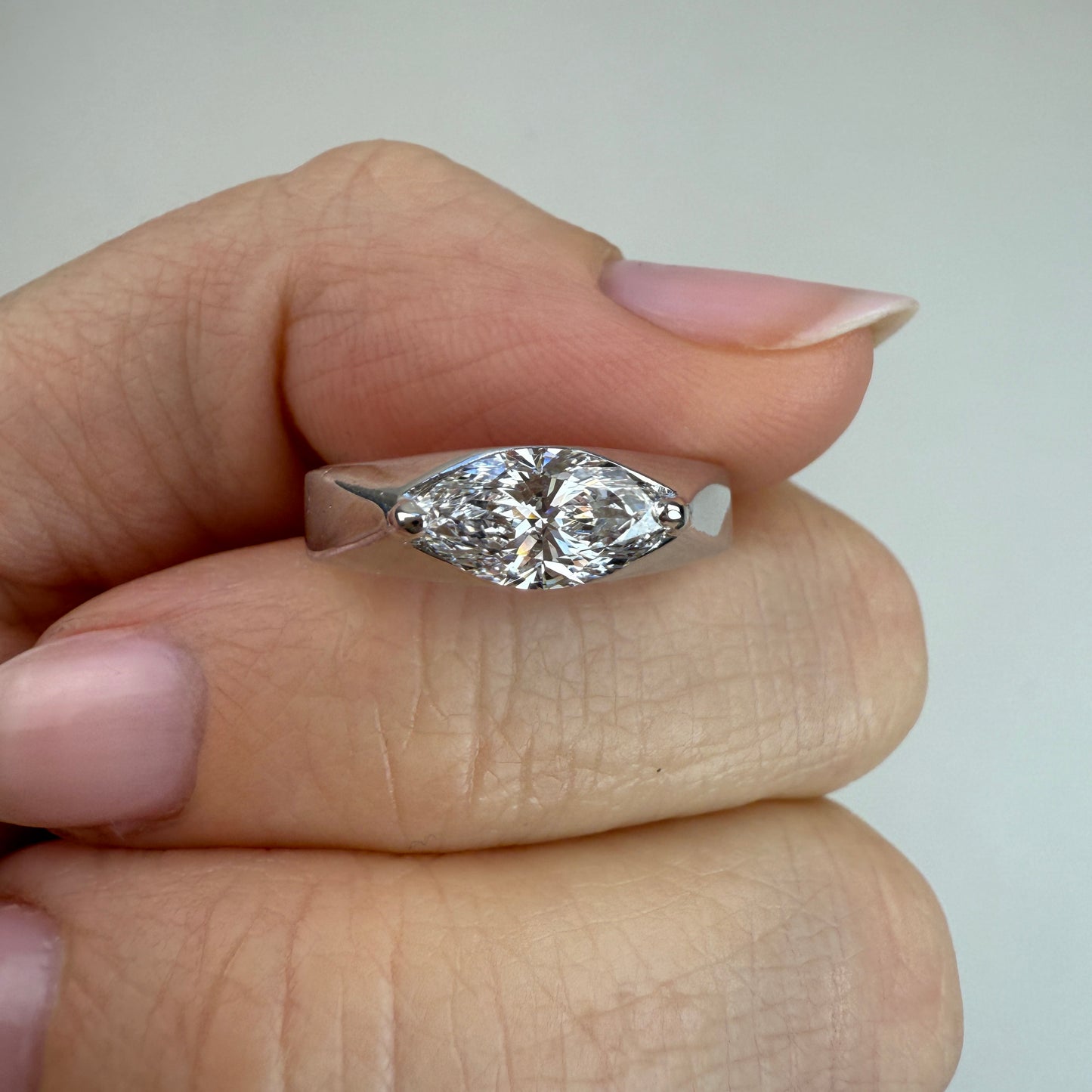 White gold and 1 carat lab marquise diamond between two fingers with off-white background.