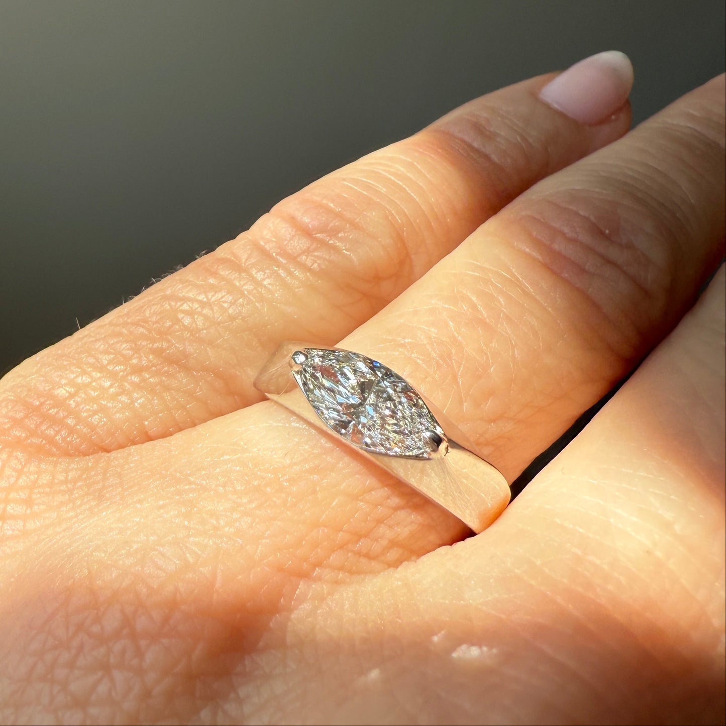 White gold and a 1-carat lab marquise diamond on the ring finger with an off-white background in the sunlight. 